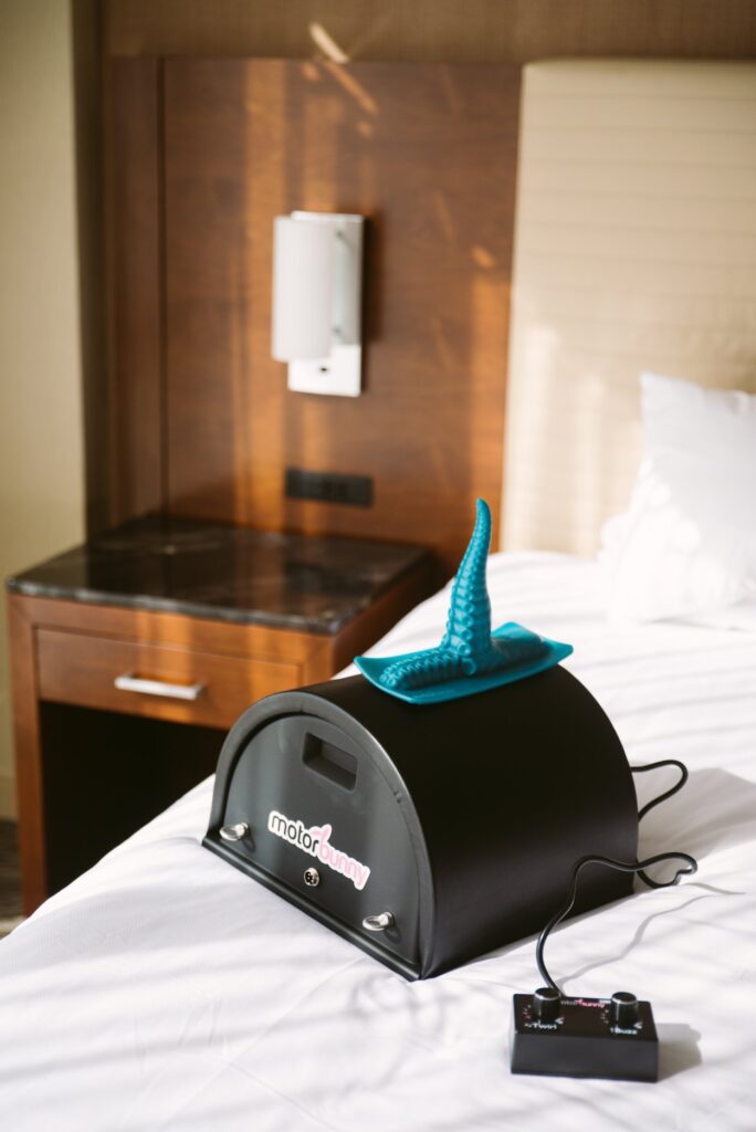 A Motorbunny sex machine sits out on a bed. The bed looks soft and welcoming. A tentacle dildo is strapped to the top of the Motorbunny machine. For my Sybian for Male? article.