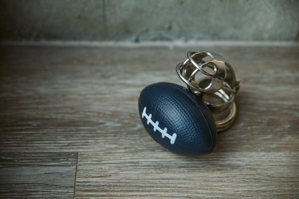 A small football-shaped stress ball sits out next to a metal chastity cage. For my femdom football scenes article.