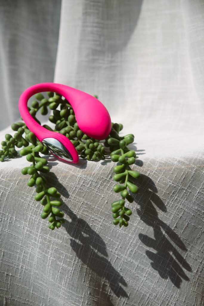 The Lovense Lush Mini is sitting on top of some greenery on top of a cheesecloth-textured backdrop. Harsh shadows fall on top of the greenery, giving long shadows that add visual interest to the shot. For my Lovense Lush Mini review.