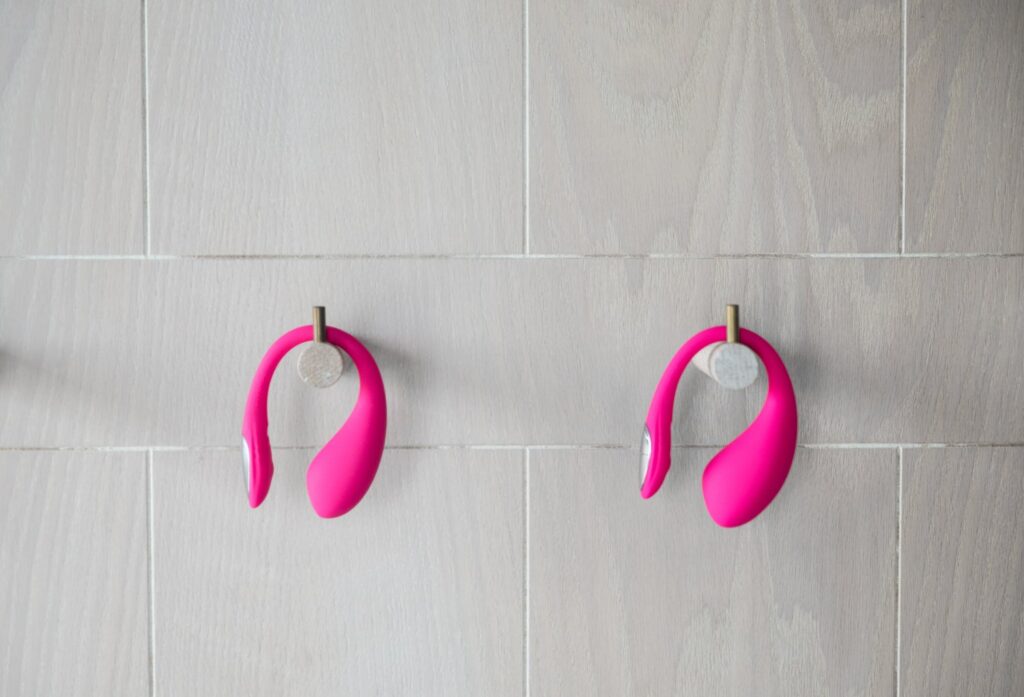 The Lovense Lush Mini and Lovense Lush 4 are hung on pegs next to one another. The Mini, which is on the left, is noticeably smaller in size. For my Lovense Lush Mini review.