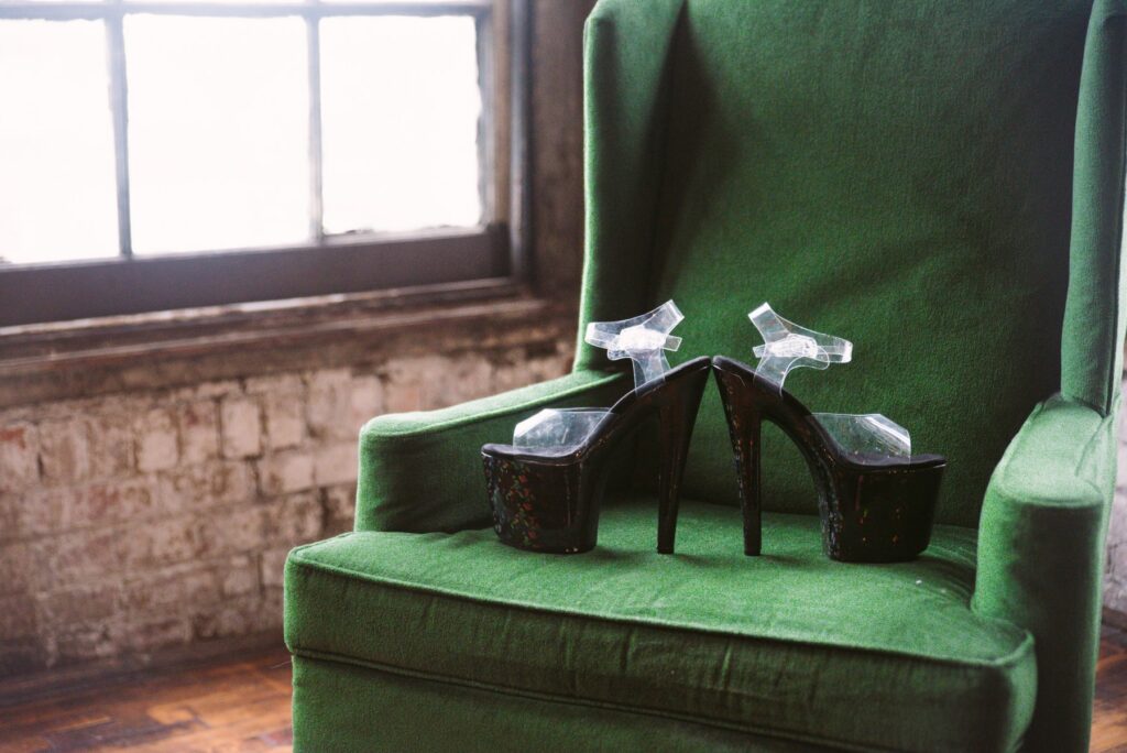 A pair of high-heeled stripper heels are sitting out on top of a green armchair. They look shiny in the nearby windowlight. For my Crossdresser High Heels Scene Ideas article.
