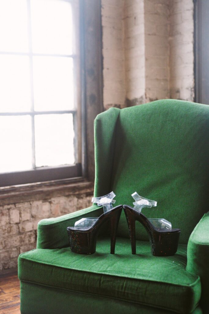 A pair of high-heeled stripper heels are sitting out on top of a green armchair. They look shiny in the nearby windowlight. For my Crossdresser High Heels Scene Ideas article.