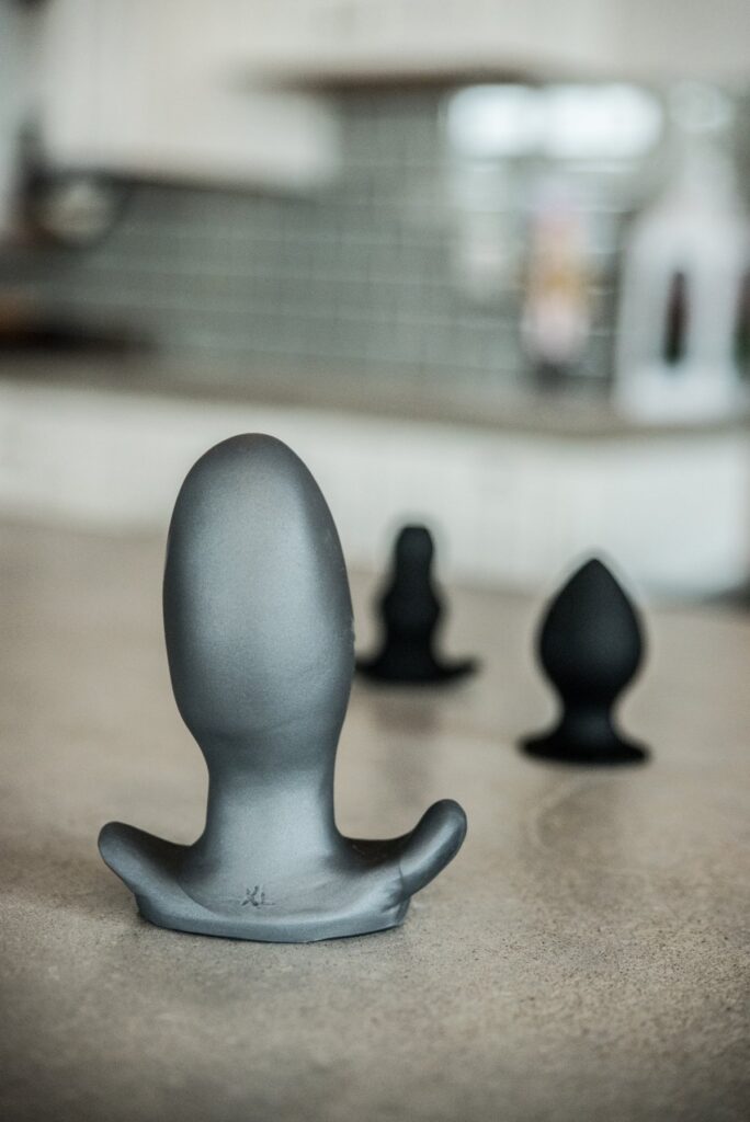 Three butt plugs are shown on a countertop. An extra-large one is shown closest to the camera with slimmer toys shown in the background. For my How to Choose the Best Toys for Anal Stretching article.