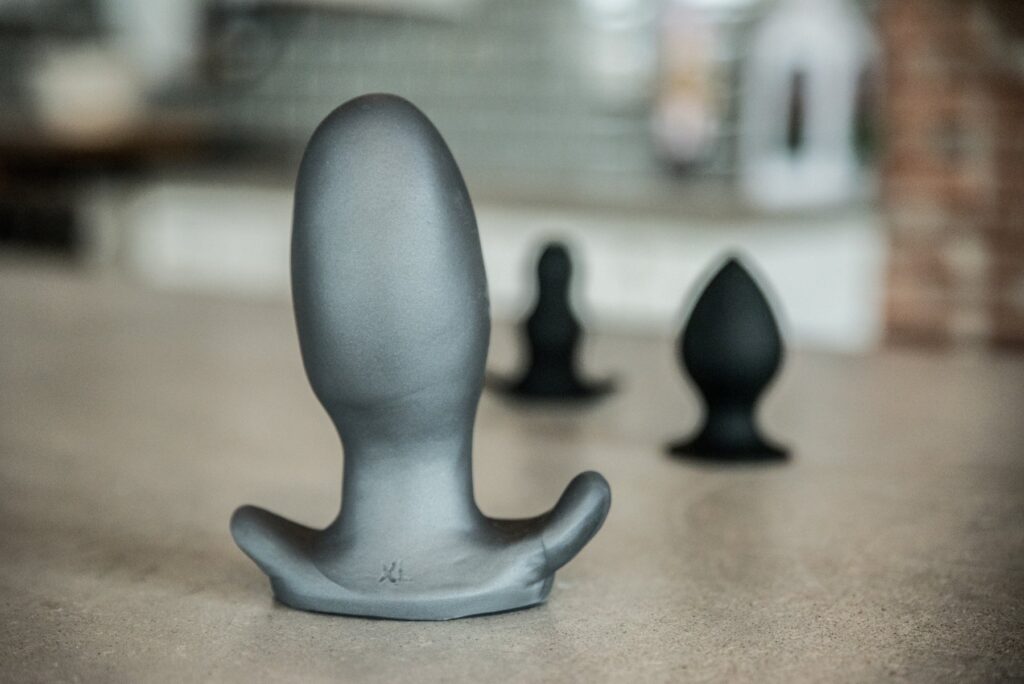 Three butt plugs are shown on a countertop. An extra-large one is shown closest to the camera with slimmer toys shown in the background. For my How to Choose the Best Toys for Anal Stretching article.