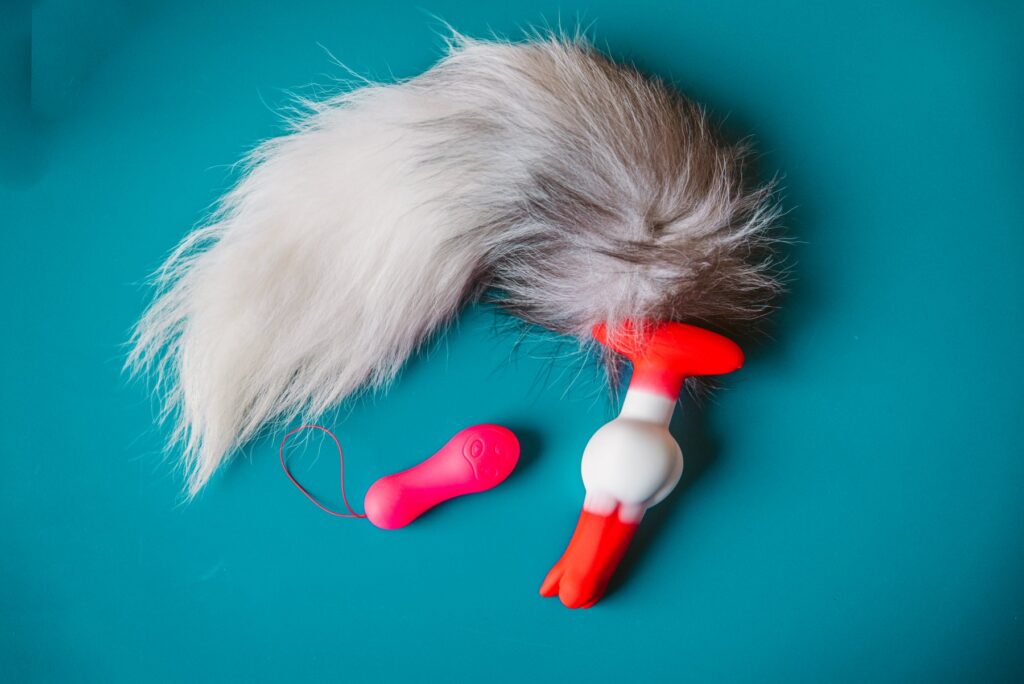 The plug, with tail attached, sits in front of a blue background. The bright pink remote sits nearby. The colors are very vibrant, and the tail looks extremely soft. For my Juliet Toys Echo review.