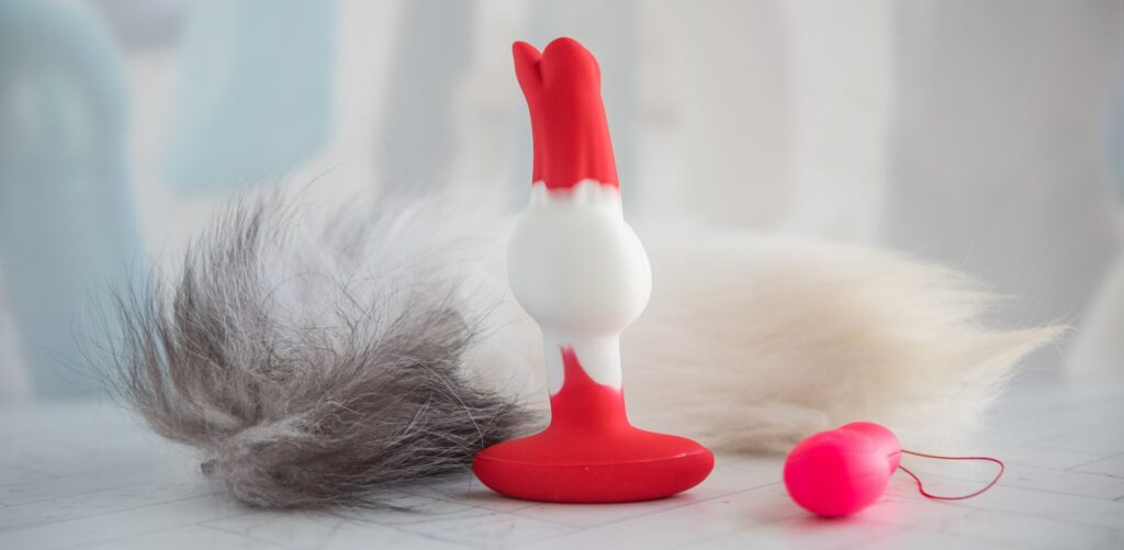 The butt plug is shown sitting upright on a flat surface. It looks long with a thick knot near the mid-point of the plug. The tail and the remote control for the vibrator sit next to the plug. For my Juliet Toys Echo review.