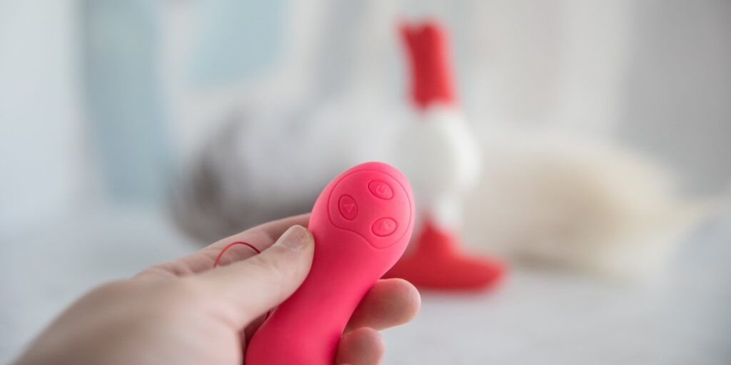 My hand holds the remote control for the sex toy pointing at it in the background. There are three buttons on the remote. A Plus, Minus, and Power button.