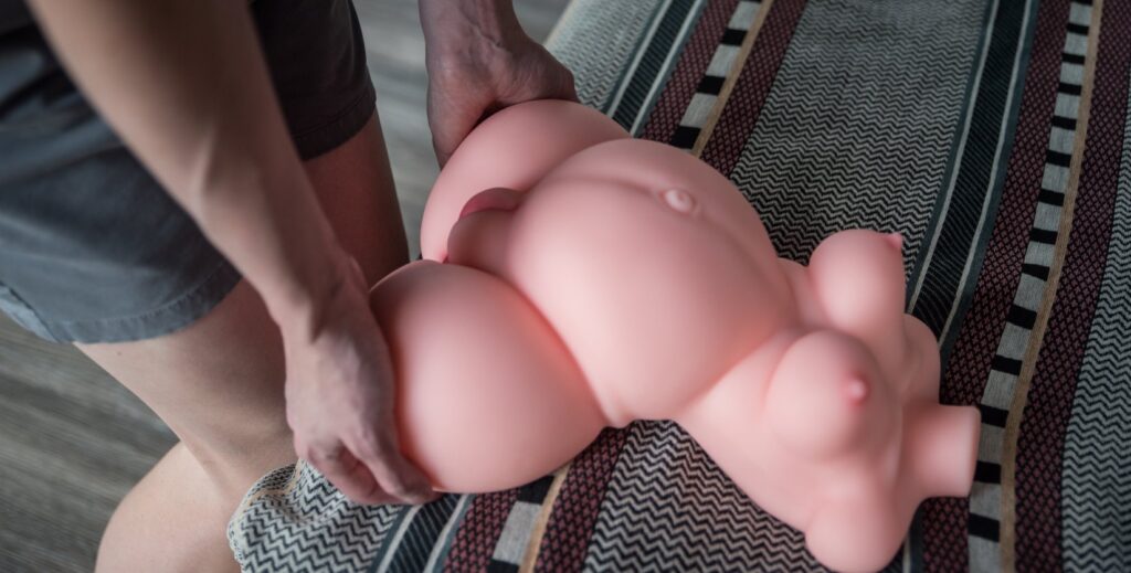 A person stands next to a bed with their hands on the thighs of the sex doll. It looks like it could be used while standing for standing sex positions.
