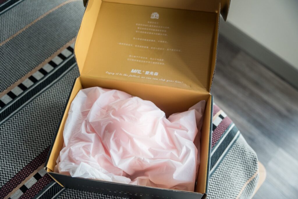 The sex doll's box is open, showcasing the sex doll inside of its packaging bag and inside of the box. This can be used for storage. The box, despite being cardboard, looks relatively sturdy.