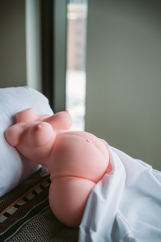 The Marissa sex doll laying on a bed, propped up by a pillow. A white sheet covers up the vulva of the doll, but the large, round pregnant tummy and the breasts are visible. For my MRLSexDoll Marissa review.