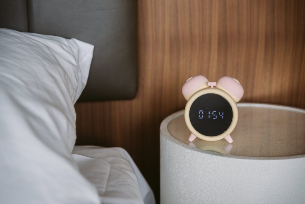 The alarm clock sits on a bedside table, brown wood shown behind it. It looks like a standard, cute alarm clock with the time digitally displayed on it. For my BeYourLover Naughty Alarm Clock review.