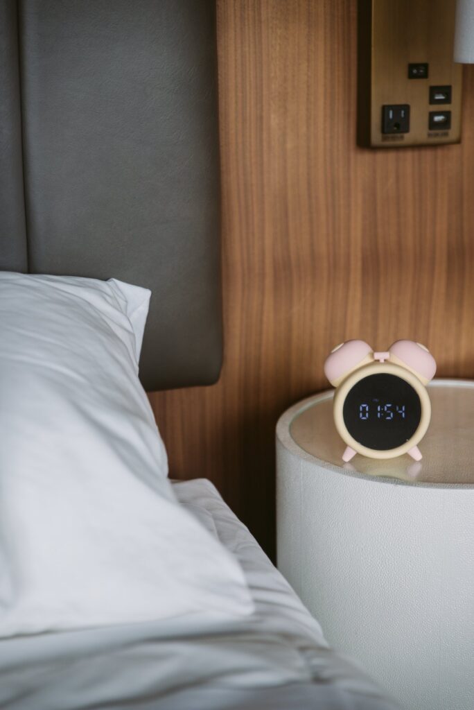 The alarm clock sits on a bedside table, brown wood shown behind it. It looks like a standard, cute alarm clock with the time digitally displayed on it. For my BeYourLover Naughty Alarm Clock review.