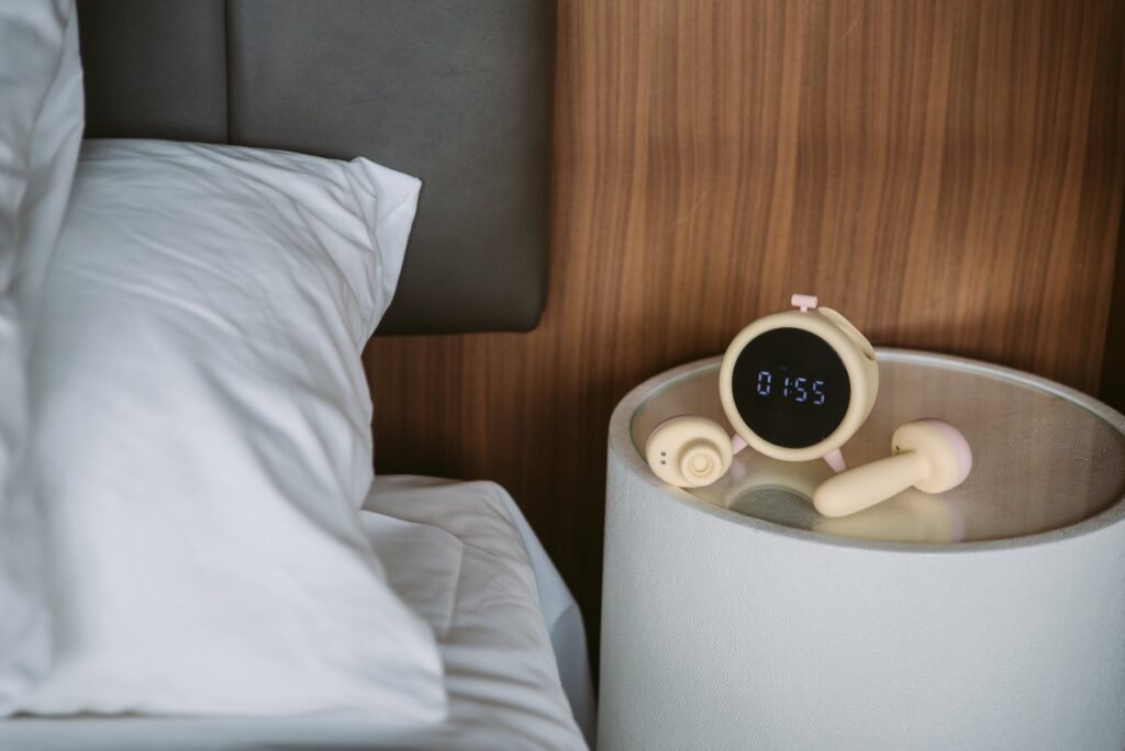 The alarm clock sits on a bedside table, brown wood shown behind it. The two sex toys are pulled out from the tops of the alarm clock, resting next to the clock. For my BeYourLover Naughty Alarm Clock review.