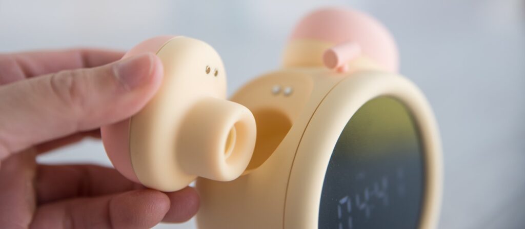 My fingers hold the air suction toy away from the base of the alarm clock. The magnetic charging port connectors on both the alarm clock itself and the air suction toy are visible, showcasing how the two snap together. For my BeYourLover Naughty Alarm Clock review.