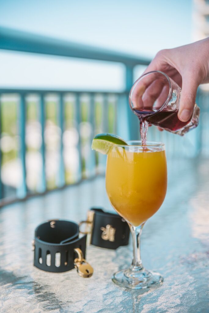 A glass of orange liquid, garnished with a sliced lime, is sitting outdoors on a glass table. Black bondage cuffs are resting next to its base. A hand, off-camera, is pour red liquid into the glass, and it mixes as it's poured. For my Femdom Cocktail Service: Everything You Need to Know article.