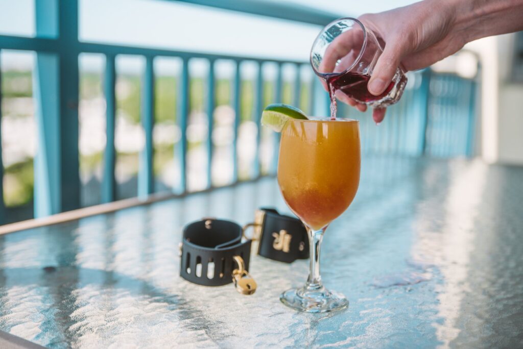 A glass of orange liquid, garnished with a sliced lime, is sitting outdoors on a glass table. Black bondage cuffs are resting next to its base. A hand, off-camera, is pour red liquid into the glass, and it mixes as it's poured. For my Femdom Cocktail Service: Everything You Need to Know article.