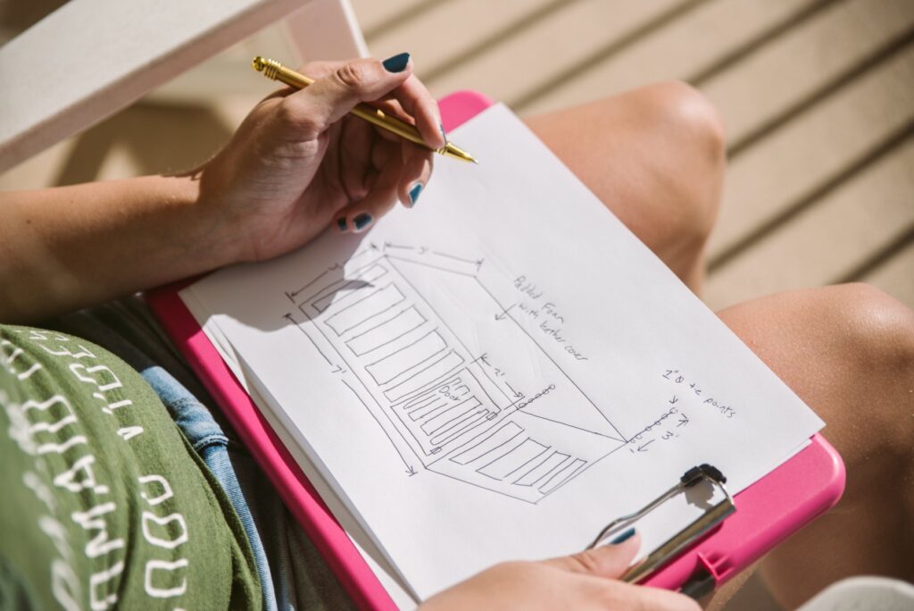 A person sits out on a rocking chair on a sunny balcony. A pink clipboard is on their lap, and they're using a pen to draw out their personalized plans for a BDSM cage, complete with measurements. For my How to Order Custom Sex Furniture article.