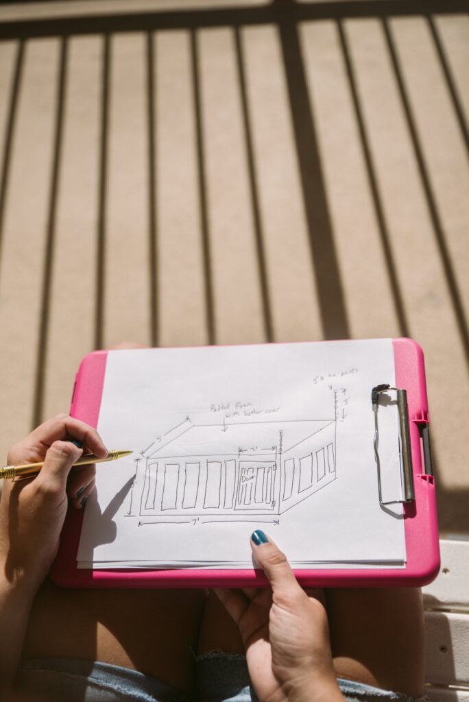 A person sits out on a rocking chair on a sunny balcony. A pink clipboard is on their lap, and they're using a pen to draw out their personalized plans for a BDSM cage, complete with measurements. For my How to Order Custom Sex Furniture article.