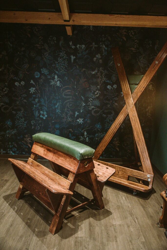 A BDSM spanking bench next to a St. Andrew's cross in a moody-looking dungeon with dark, floral wallpaper. For my BDSM safe word and safe action comprehensive article.