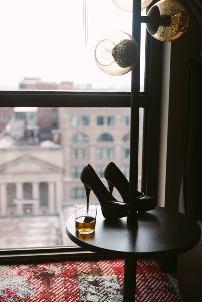 A pair of black heels is backlit next to a window. There's a cup of bourbon-like liquid in a two fingers glass. For my how to be a dom woman article.