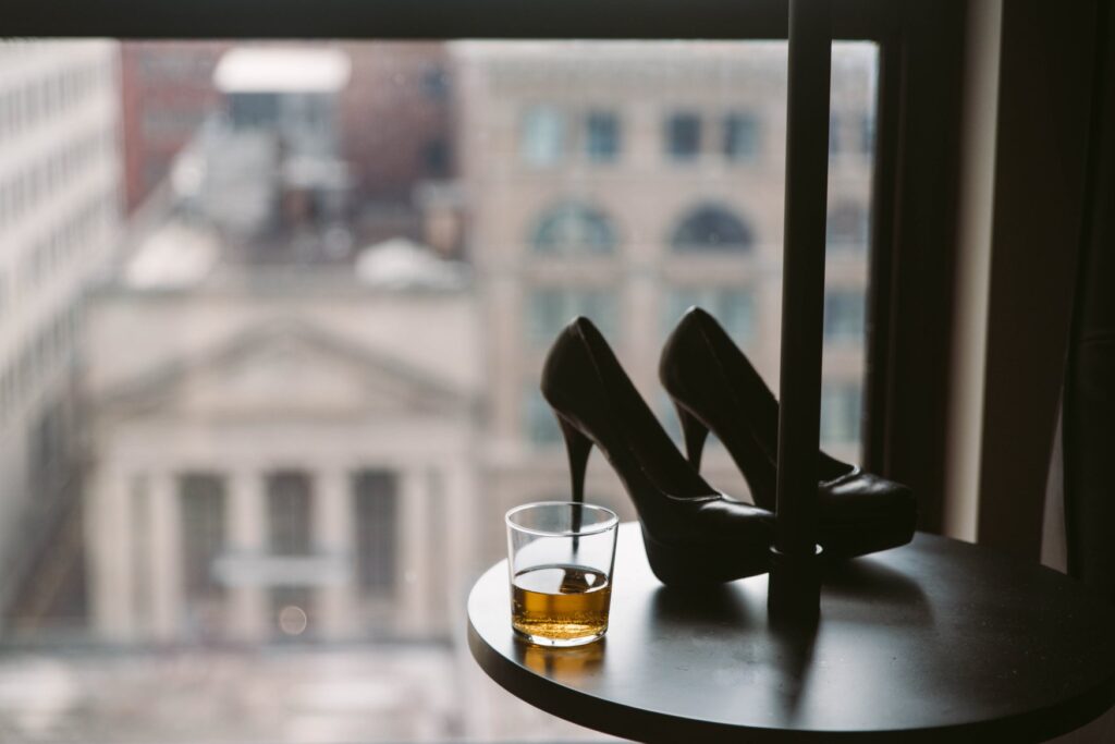 A pair of black heels is backlit next to a window. There's a cup of bourbon-like liquid in a two fingers glass. For my how to be a dom woman article.