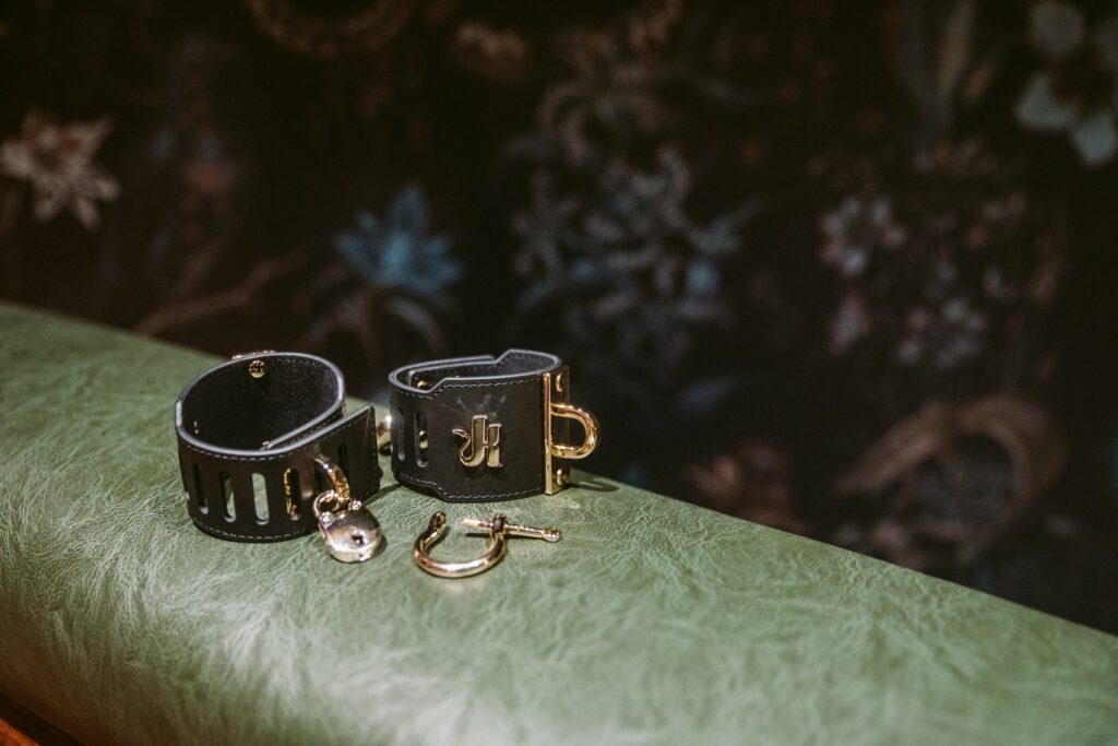 The black and gold cuffs sit out on a green spanking bench in front of a wooden St. Andrew's cross. The "K" golden logo is shiny and visible. For my Kink.com Gold Eclipse Wrist Cuff set review.