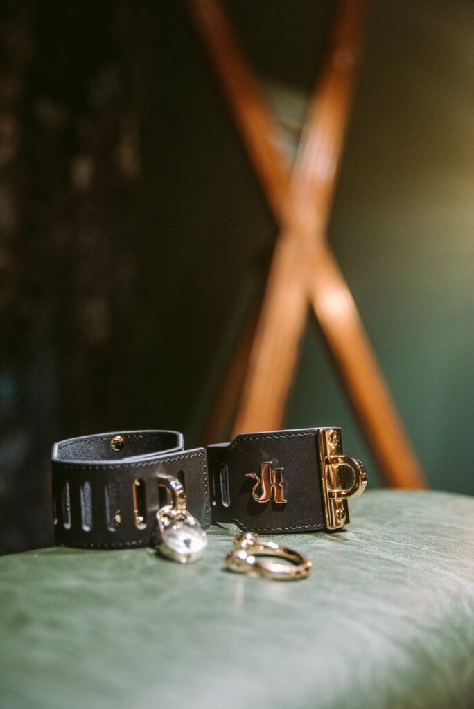 The black and gold cuffs sit out on a green spanking bench in front of a wooden St. Andrew's cross. The "K" golden logo is shiny and visible. For my Kink.com Gold Eclipse Wrist Cuff set review.