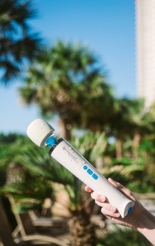 A hand holds the Magic Wand Waterproof up in the air outdoors, with a beach and palm trees in the background. For my Magic Wand Waterproof review.
