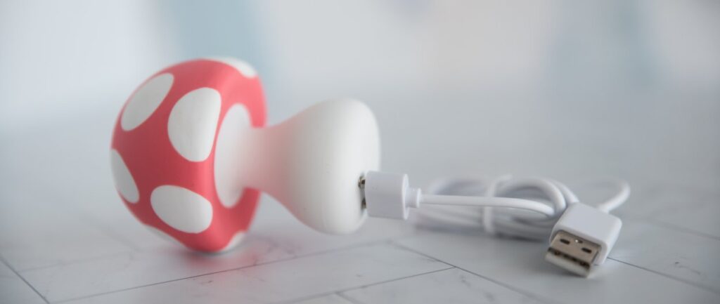 The Mushroom Vibez toy lays on its side. The magnetic charger is connected to the base of the vibrator, adhered to the two magnetic charging ports on the base. For my Mushroom Vibez review.