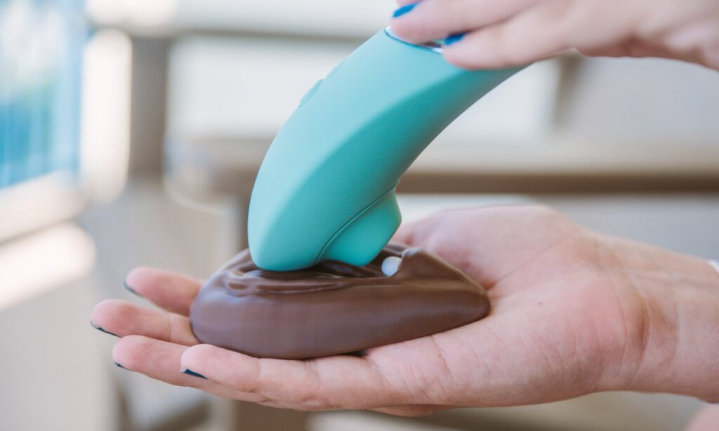 A person holds a vulva in their hands. They have positioned the Womanizer Next, an air suction vibrator, at an under-the-clit angle, enveloping the clit in the tip of the toy. For my How to Use a Clit Suction Toy article.