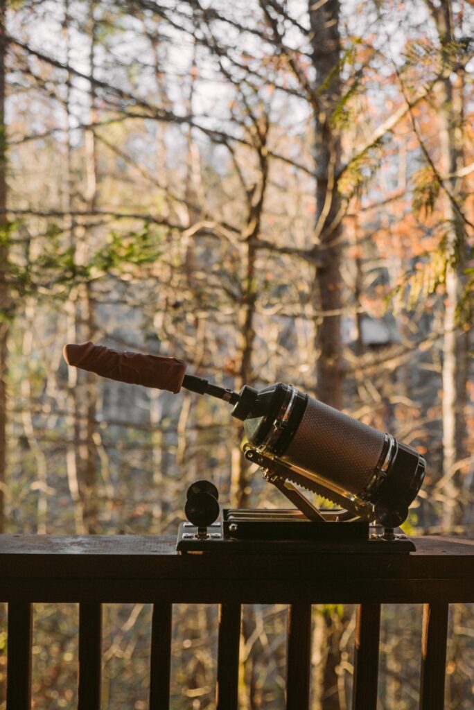 A sex machine sits out on top of a balcony ledge. The machine has a dildo on it. There are autumnal trees in the background. For my article about Are Sex Machines Worth It?