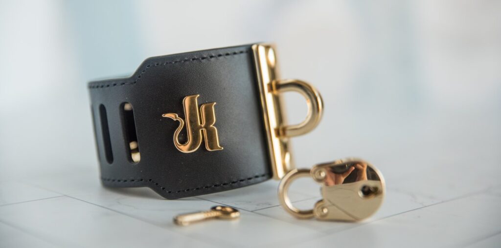 The cuff with the iconic Kink.com K emblem towards the camera. It's a nice, strong, shiny metal that color matches the rest of the hardware. The color-matching lock and key are shown in the foreground, equally reflective.