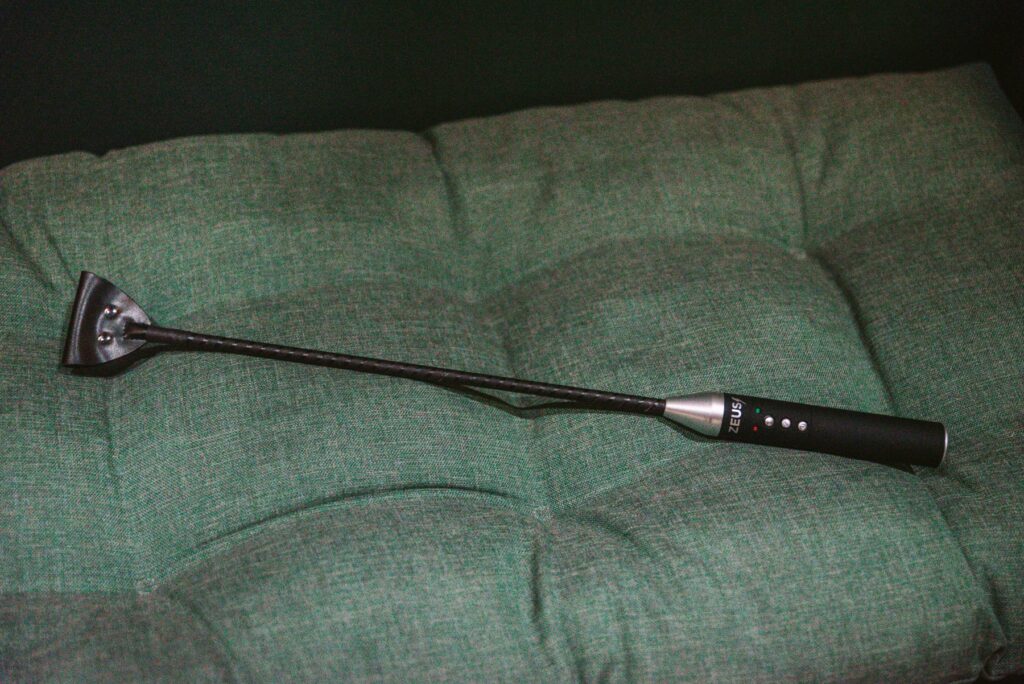 The Zeus e-Stim Riding Crop lies on top of a green, cushioned bench. It's broadly lit, showcasing the full length of the crop.
