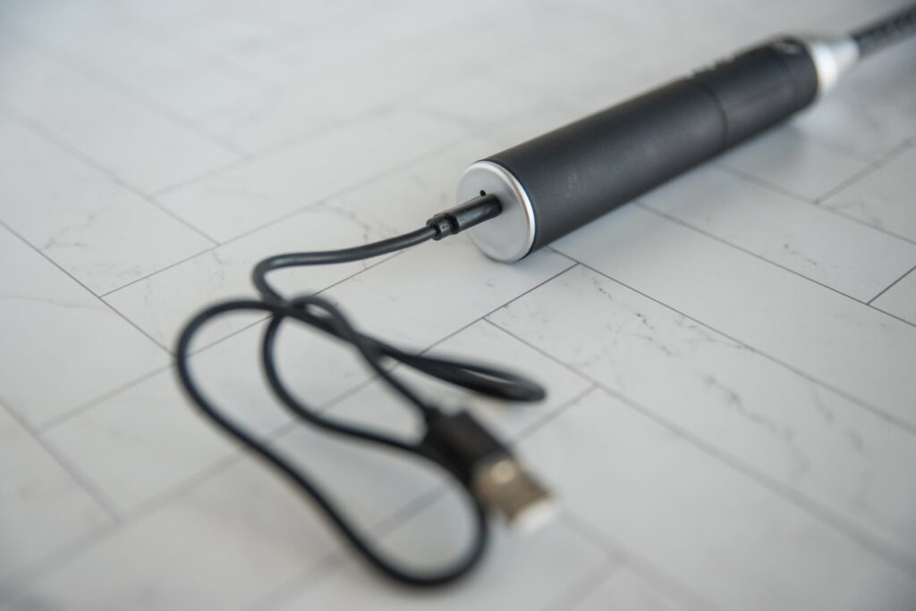 Close-up on the base of the electrified riding crop. The charging cable is plugged into the open charging port. The other end of the cable is a USB end. For my Dr. Sado e-Stim Crop review.