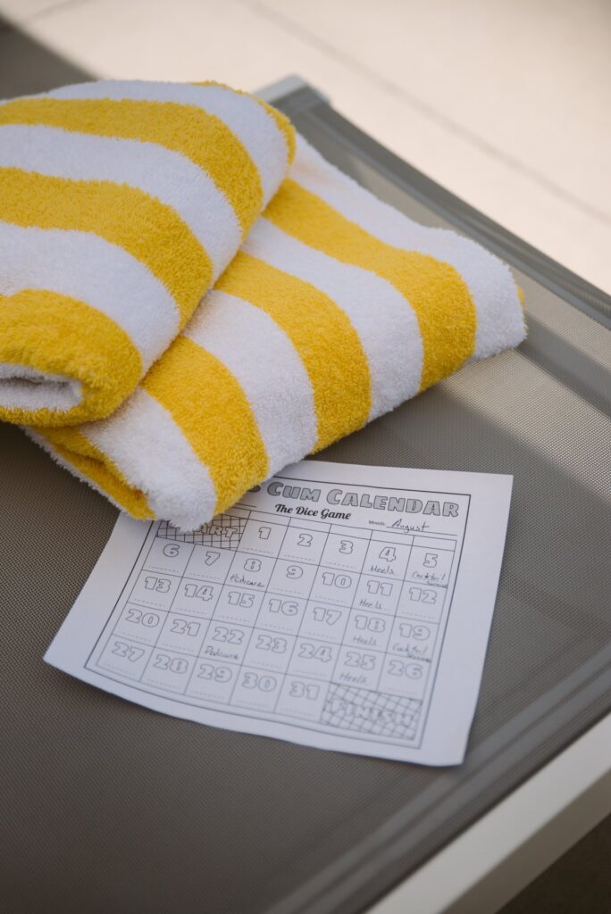 A "Cum Calendar" print out is sitting out on a beach chair, next to a beach towel. Some of it is filled in. For my how to build your own BDSM training plan.