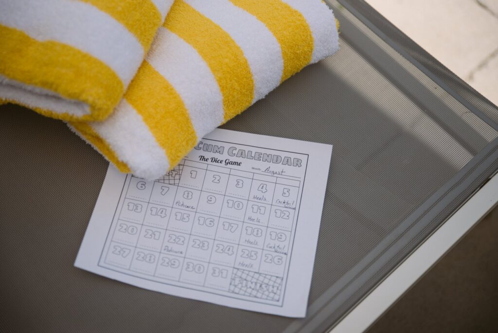 A "Cum Calendar" print out is sitting out on a beach chair, next to a beach towel. Some of it is filled in. For my how to build your own BDSM training plan.