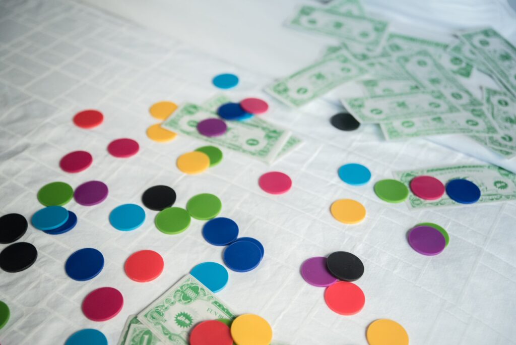 A lot of colorful flat game tokens and fake cash bills are spread out across the surface of a bed's white bedding. It looks very fun.