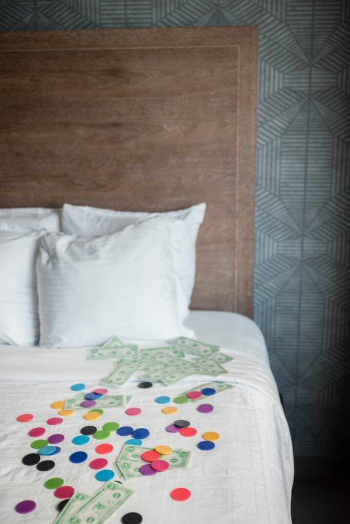 A lot of colorful flat game tokens and fake cash bills are spread out across the surface of a bed's white bedding. It looks very fun.