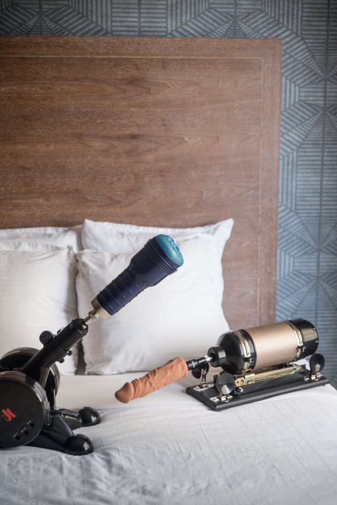 Two sex machines are sitting out on a bed. One sex machine is thrusting a Fleshlight back and forth while the other has a dildo that it can thrust back and forth. For my DIY sex robot BDSM Scenes, Step-by-Step plan.