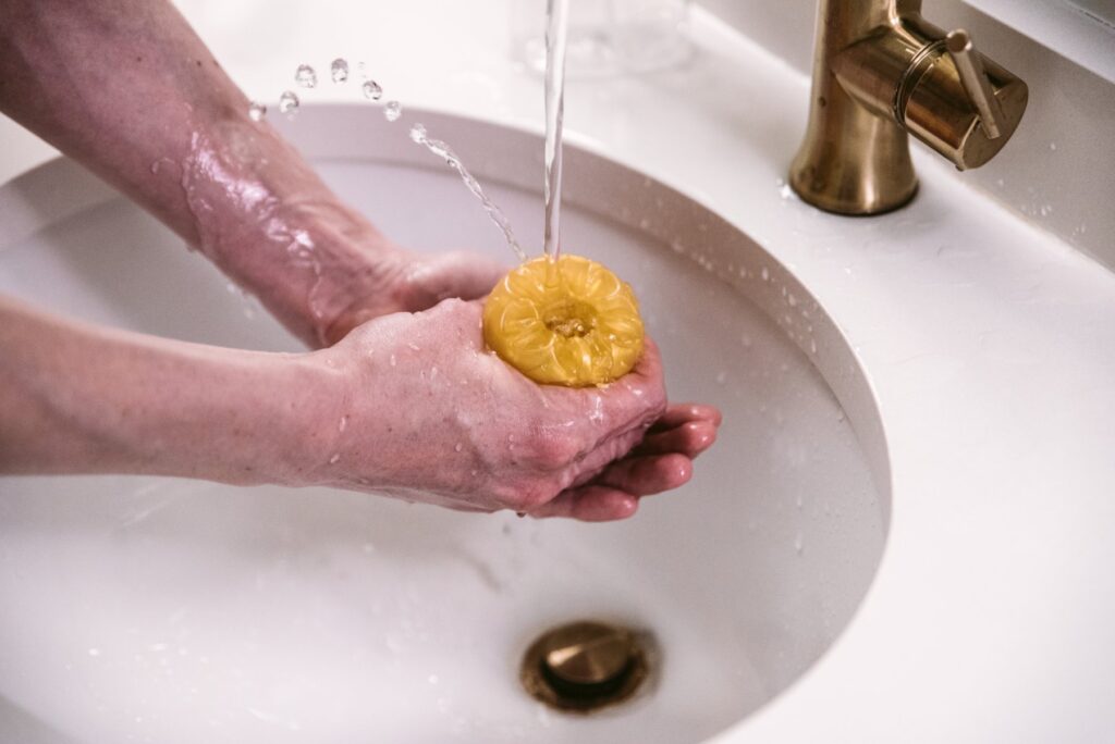 A stroker is held open underneath a sink spigot of running water. Another hand blocks the other open side of the stroker, causing all of the water to spurt back out the top. For my How to Clean a Pocket Pussy article.