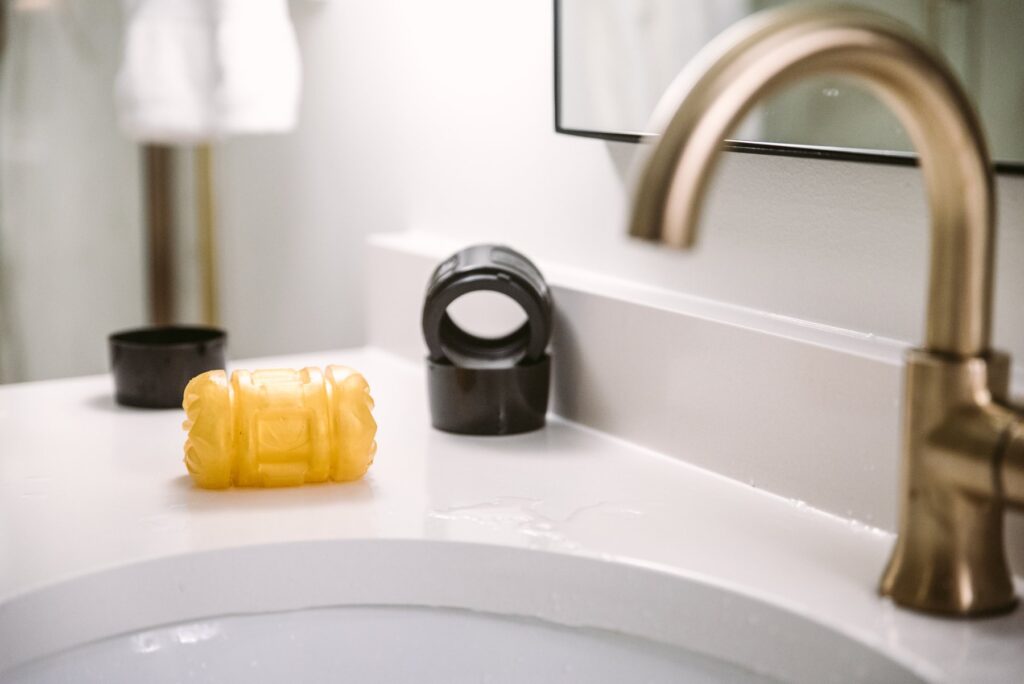 A penis stroker sits out on the side of a sink. The case of the sex toy is sitting nearby, but the stroker itself is sitting out by itself to allow the toy to dry.