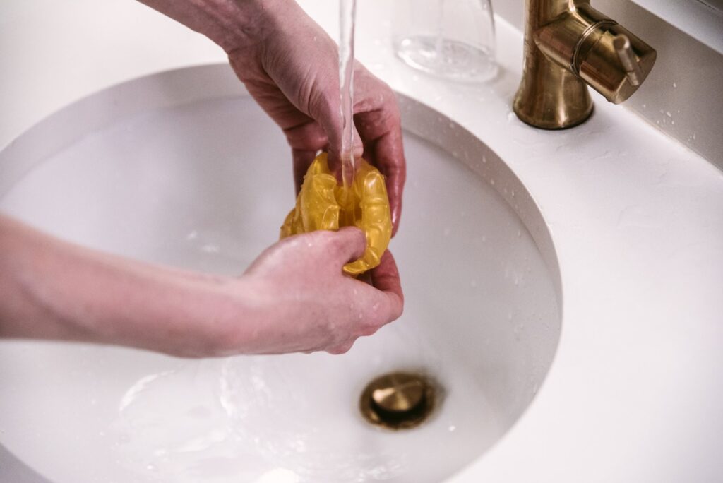 A stroker is held open underneath a sink spigot of running water. The water is running right into the open chamber of the stroker. For my How to Clean a Pocket Pussy article.
