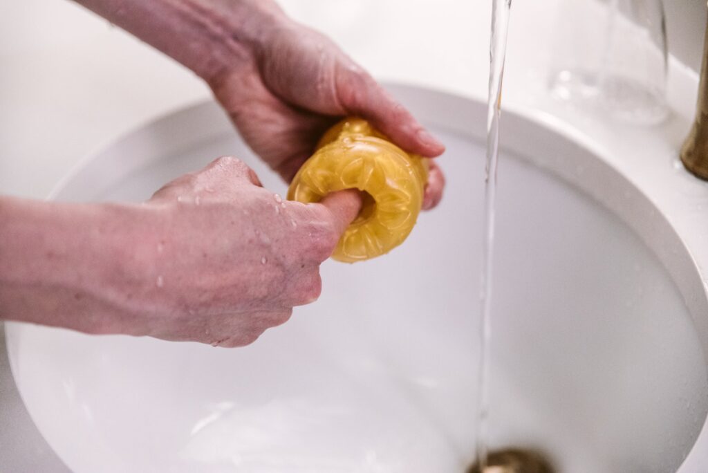 A stroker is held open underneath a sink spigot of running water. A hand slides fingers inside of the stroker, trying ti dislodge anything inside and make sure the entire interior was covered in water. For my How to Clean a Pocket Pussy article.
