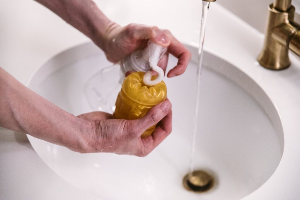A stroker is held open underneath a sink spigot of running water. A hand squirts mild, un-oiled hand soap onto the orifice of the stroker. For my How to Clean a Pocket Pussy article.