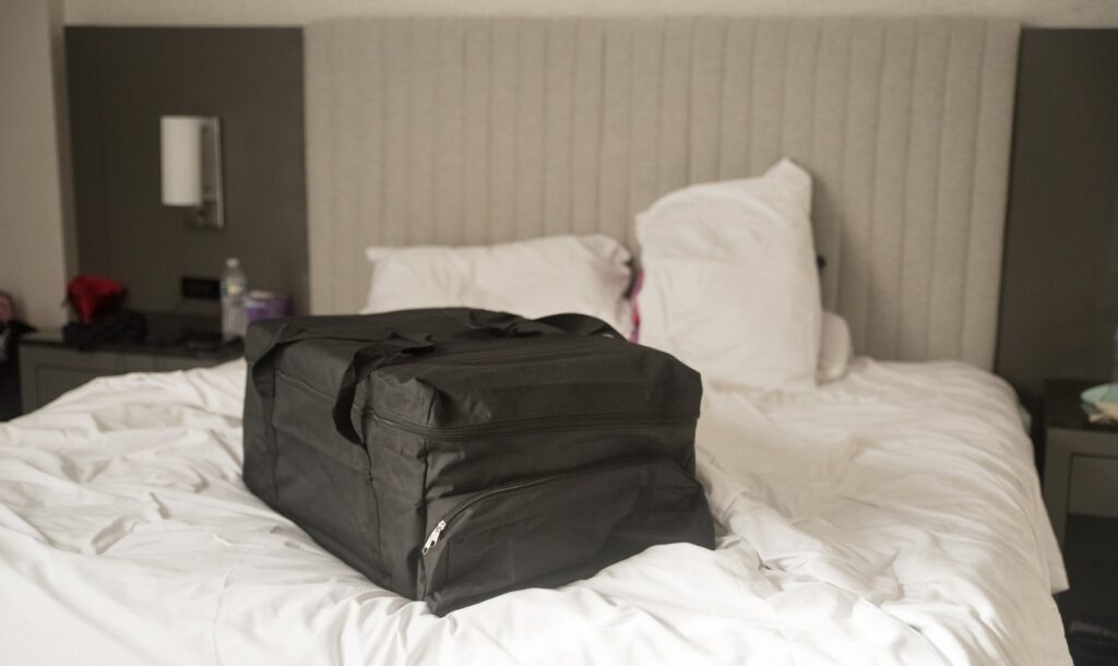 The black storage bag that is included with the sex doll. It is sitting on a bed and looks very large.