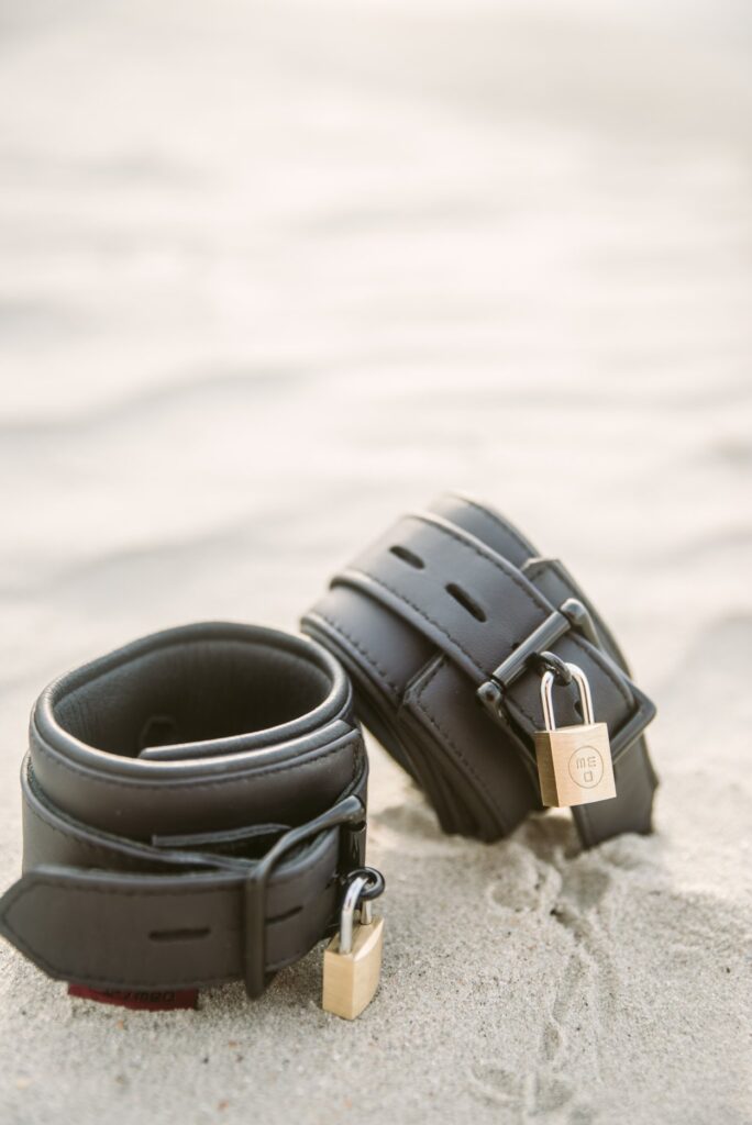 Two neoprene, waterproof cuffs set out on the sand. They both have metal locks, locking the cuffs into their fastened position. For my Tips for Beach Bondage article.