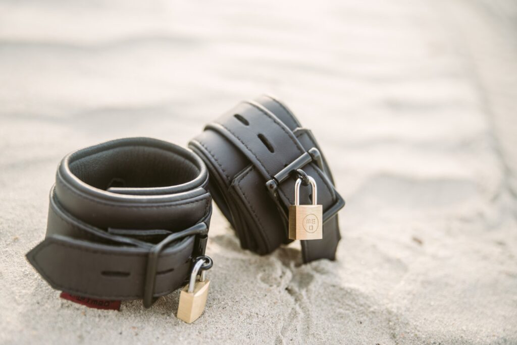 Two neoprene, waterproof cuffs set out on the sand. They both have metal locks, locking the cuffs into their fastened position. For my Tips for Beach Bondage article.