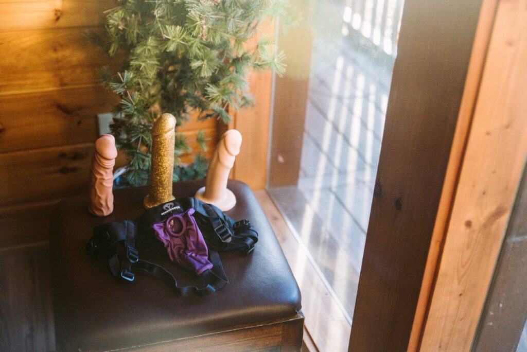 A footrest sitting next to a window with golden hour light cascading through the window. The footrest has a strap-on harness and three dildos sitting on it. For my How to Host a Femdom Pegging Party.