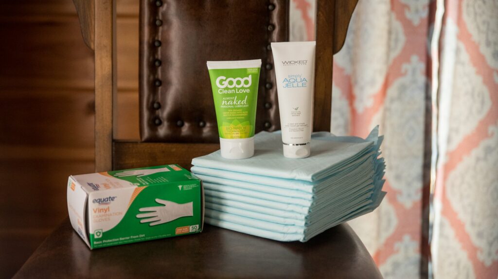 Items sitting out on a chair. There are two bottles of lube, a stack of puppy pads, and a box of disposable hand gloves.