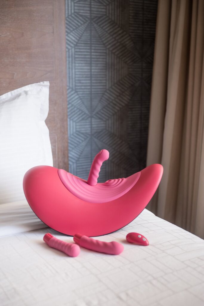 The VVD Rider Desire machine sits out on the edge of a bed. One of its attachments is in it, but the other two attachments and the remote sit next to it. For my VVD Rider Desire review.